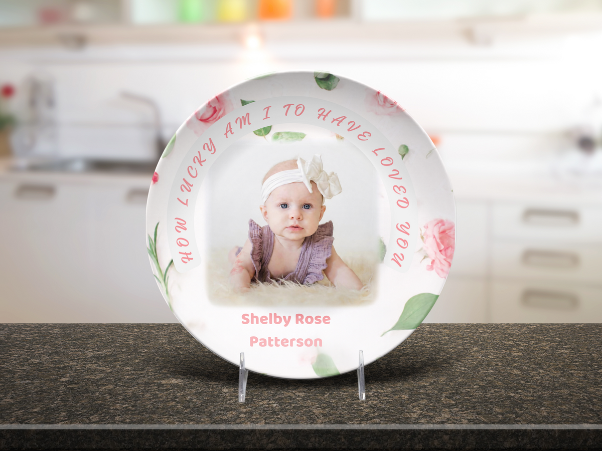 A personalized ceramic baby keepsake plate displayed on a clear stand on a dark countertop, with a blurred kitchen background. The white plate features pink floral designs, green leaves, and pink text reading 'HOW LUCKY AM I TO HAVE LOVED YOU' and 'Shelby Rose Patterson'.  Stand is optional.