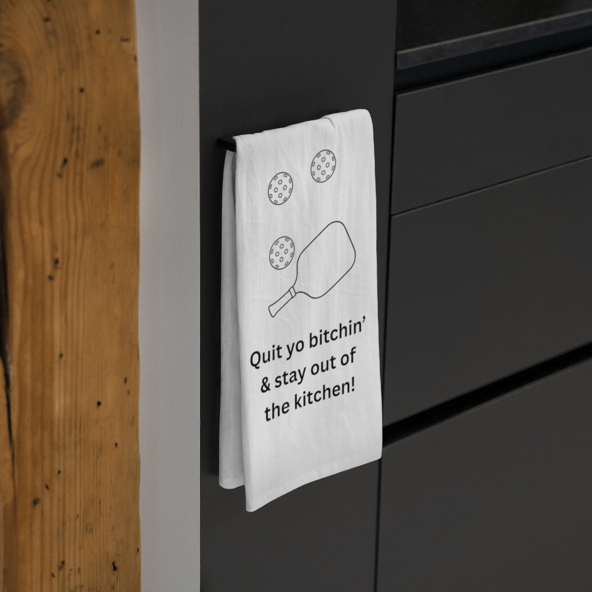 White tea towel with a pickleball design hanging on a black cabinet. 